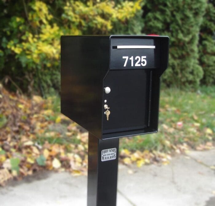 Tall, square mailbox over sidewalk, with black paint and white numbers with a small key placed in the lock